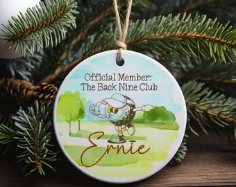 May include: Round ceramic ornament with a golf-themed cartoon character, "Ernie," on a green and blue background. The ornament has the text "Official Member: The Back Nine Club."