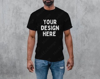 Puede incluir: Una camiseta negra con el texto "YOUR DESIGN HERE" impreso en blanco en la parte delantera. La camiseta la lleva puesta una persona de piel oscura y va combinada con unos vaqueros azules.