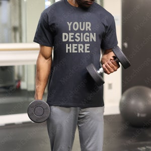 May include: A person wearing a black t-shirt with the text "YOUR DESIGN HERE" printed on the front. The person is lifting a dumbbell with their right hand. The person is in a gym setting.