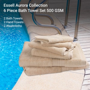 May include: A set of six beige bath towels, including two bath towels, two hand towels, and two washcloths. The towels are stacked on a wooden surface near a swimming pool. The towels are made from 500 GSM cotton and are part of the Essell Aurora Collection.