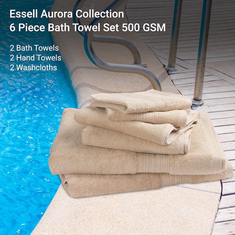 May include: A set of six beige bath towels, including two bath towels, two hand towels, and two washcloths. The towels are stacked on a wooden surface near a swimming pool. The towels are made from 500 GSM cotton and are part of the Essell Aurora Collection.