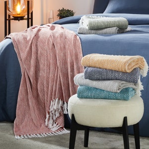 May include: A stack of five throw blankets with a herringbone pattern in various colors, including red, yellow, teal, gray, and green. The blankets are folded and stacked on a white stool with black legs.