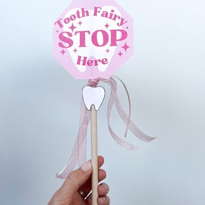 May include: Pink tooth fairy stop sign with the words "Tooth Fairy Stop Here" and a white tooth graphic. The sign is attached to a wooden stick with a pink ribbon.