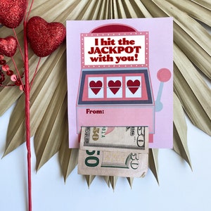 Pu&ograve; includere: Un biglietto rosa di San Valentino con un design a slot machine e la scritta "I hit the JACKPOT with you!". La slot machine ha tre simboli a forma di cuore. Sono visibili due banconote da cinquanta dollari. Cuori rossi decorativi e una foglia di palma secca sullo sfondo.