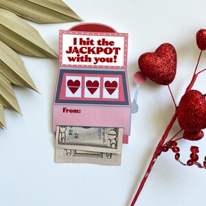 Pu&ograve; includere: Un biglietto a forma di slot machine rosa e grigio con la scritta "I hit the JACKPOT with you!" e tre simboli di cuore. Il biglietto ha uno scomparto per contanti, con due banconote visibili. Una decorazione a forma di cuore rosso glitterato &egrave; a destra.