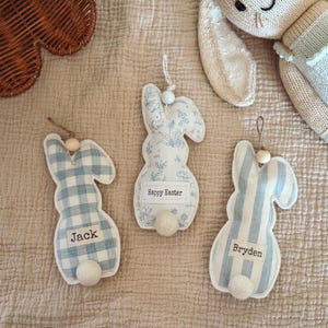May include: Three Easter bunny decorations in blue and white patterns, with a knitted bunny and a woven basket in the background. One ornament says "Happy Easter", and the others are personalised with names. Each bunny has a white pom-pom tail.