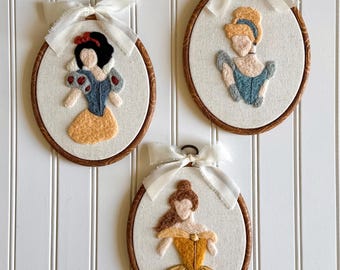 May include: Three oval-shaped wall hangings featuring felted princess designs. Each piece has a wooden frame, a white background, and a white ribbon bow at the top. The princesses are Snow White, Cinderella, and Belle, each in their iconic outfits.