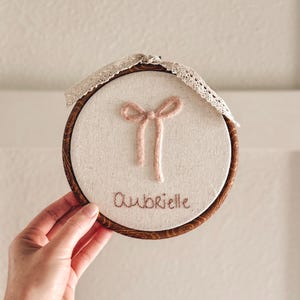 May include: An embroidered hoop with a light fabric background. It features a pink bow and the name "Aubrielle" stitched in brown thread. The wooden hoop is trimmed with decorative lace. The hoop is round.