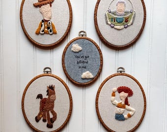 May include: Five oval wooden embroidery hoops containing fabric art. Four hoops feature embroidered characters including a cowboy, a space ranger, a horse, and a cowgirl. The center hoop features a blue background with white clouds and the text You've got a friend in me.