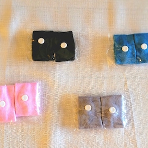 May include: Five pairs of wristbands in various colors: black, blue, pink, and gray. Each pair is packaged in clear plastic and features two white buttons. The wristbands appear to be made of a textured fabric.