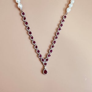 Può includere: Una collana d'argento con un ciondolo a forma di lacrima in rubino circondato da diamanti più piccoli. La collana è composta da una catena di perle e pietre di rubino.