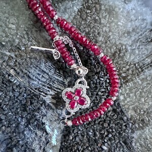 May include: A red ruby gemstone beaded bracelet with a silver flower charm. The charm has a small ruby gemstone in the center and is surrounded by smaller white gemstones.