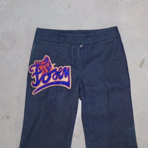 May include: Dark blue denim trousers with a decorative patch on the left leg. The patch features the word "Foxy" in a stylized font, with a red and orange design outlined in blue.