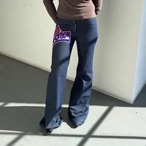 May include: Dark wash denim jeans with a flared leg. The jeans feature a patch on the front with the word "FOREM" in a blue and orange design. The person is wearing a brown long-sleeve shirt and black shoes.