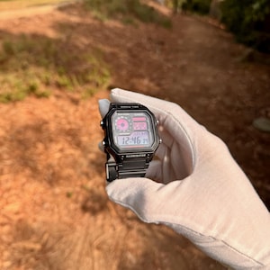 May include: A black digital wristwatch with a pink display. The watch has a black band and a square face. The watch face displays the time, date, and other information. The watch is being held in a white gloved hand.