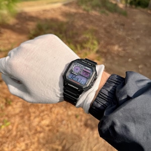 May include: A black Casio digital watch with a black strap. The watch face shows the time, date, and day of the week. The watch also has a backlight function.