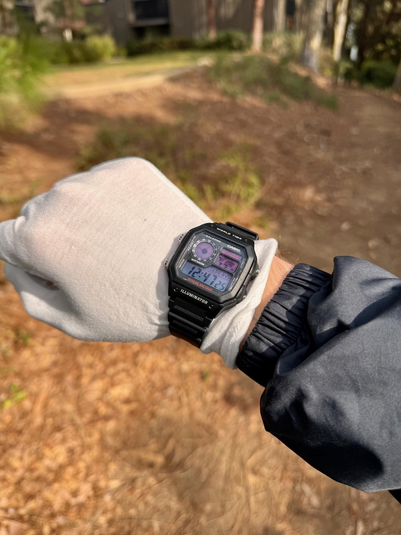 May include: A black Casio digital watch with a black strap. The watch face shows the time, date, and day of the week. The watch also has a backlight function.