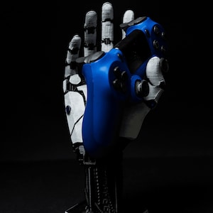 May include: A 3D printed robotic hand holding a blue video game controller. The hand is white and gray, and the controller has black buttons.