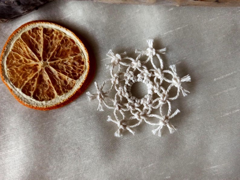 May include: A white crocheted snowflake ornament on a beige fabric background. The snowflake has a circular centre and six points.