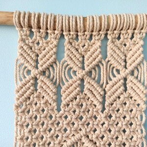 May include: A beige macrame wall hanging with a geometric pattern. The hanging is attached to a wooden dowel rod.