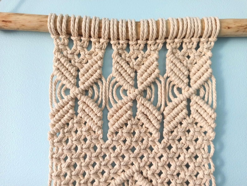 May include: A beige macrame wall hanging with a geometric pattern. The hanging is attached to a wooden dowel rod.