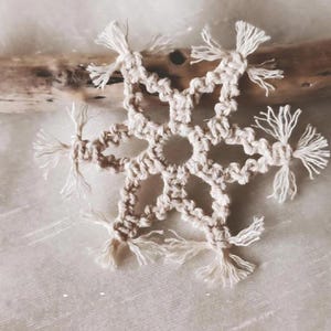 May include: A white crocheted snowflake ornament with fringe details. The snowflake is made with natural cotton cord and has a delicate, handmade look.