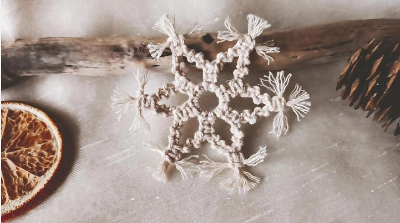 May include: A white crocheted snowflake ornament with fringe details. The snowflake is made with natural cotton cord and has a delicate, handmade look.