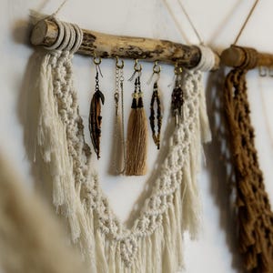 May include: A macrame earring holder with a natural wood branch and hanging earrings. The macrame is off-white and cream-colored, with a fringe design. The earrings include feathers, tassels, and beaded designs.
