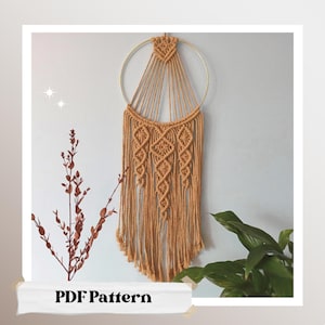 Puede incluir: Un tapiz de macramé de pared con un aro dorado y un patrón geométrico. El tapiz está hecho con cordón de color natural y tiene una franja en la parte inferior. El texto "PDF Pattern" es visible en la parte inferior de la imagen.