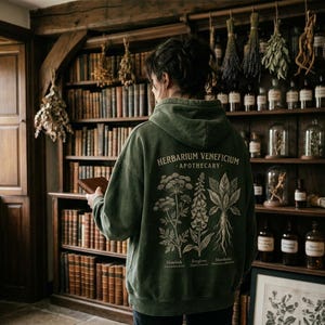May include: An olive green hooded sweatshirt featuring a back graphic with the text "HERBARIUM VENEFICIUM APOTHECARY" above botanical illustrations of Hemlock, Foxglove, and Mandrake plants with their respective scientific names printed below.