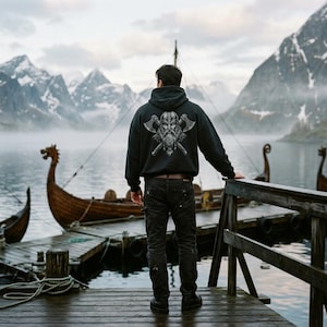 May include: A black hooded sweatshirt featuring a detailed graphic on the back of a bearded warrior wearing a helmet with two crossed axes behind the head. The hoodie has long sleeves, a drawstring hood, and a ribbed hem, worn with dark trousers.