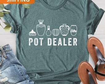 May include: Teal blue t-shirt with a white graphic of a row of potted plants and the text "POT DEALER".