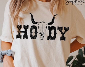 May include: A white t-shirt with a black distressed graphic of a cow skull and the word "Howdy" printed on it.
