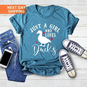 May include: A teal blue t-shirt with the text 'Just a girl who loves ducks' in white lettering with red hearts. The shirt is laid out on a woodgrain surface with a pair of blue jeans and a pair of blue converse shoes.