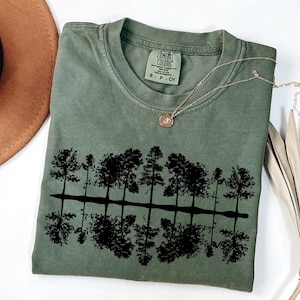 May include: A green t-shirt with a black silhouette of trees reflected in water. The shirt has the text "COMFORT COLORS" on the tag.