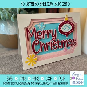 May include: A white card with a 3D layered shadow box design. The box features a blue background with the text "Merry Christmas" in red lettering. A red ornament with a white snowflake design is on the right side of the card. The card is on a wooden surface.