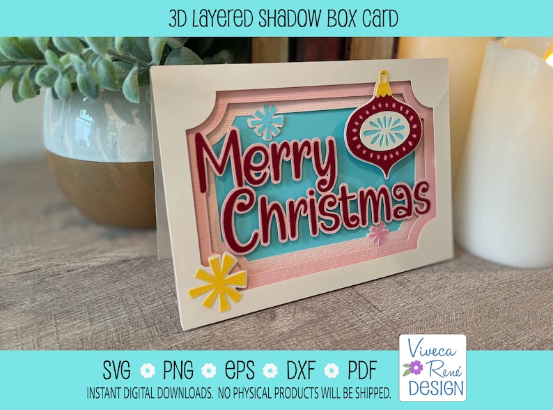 May include: A white card with a 3D layered shadow box design. The box features a blue background with the text "Merry Christmas" in red lettering. A red ornament with a white snowflake design is on the right side of the card. The card is on a wooden surface.