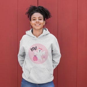 Puede incluir: Una sudadera gris con un gr&aacute;fico circular rosa que presenta un fantasma blanco con un lazo rosa, una calabaza y el texto "BOO!"