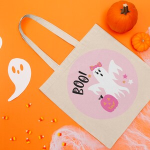 Puede incluir: Una bolsa de lona blanca con un dise&ntilde;o circular rosa que presenta un fantasma sosteniendo una calabaza y la palabra "BOO!" en negro. La bolsa tiene una correa blanca y est&aacute; sobre un fondo naranja brillante con recortes de fantasmas blancos, ma&iacute;z dulce y telara&ntilde;as blancas.