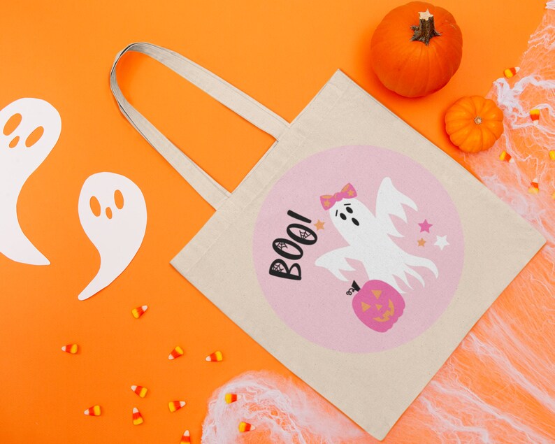 Puede incluir: Una bolsa de lona blanca con un dise&ntilde;o circular rosa que presenta un fantasma sosteniendo una calabaza y la palabra "BOO!" en negro. La bolsa tiene una correa blanca y est&aacute; sobre un fondo naranja brillante con recortes de fantasmas blancos, ma&iacute;z dulce y telara&ntilde;as blancas.