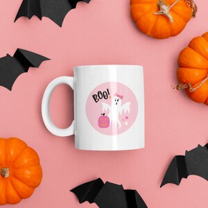 Puede incluir: Taza de cer&aacute;mica blanca con un dise&ntilde;o circular rosa que presenta un fantasma con un lazo rosa, una calabaza y la palabra "BOO!" sobre un fondo rosa. La taza est&aacute; rodeada de murci&eacute;lagos negros y calabazas naranjas sobre un fondo rosa.