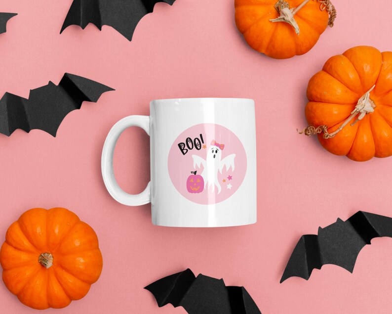 Puede incluir: Taza de cer&aacute;mica blanca con un dise&ntilde;o circular rosa que presenta un fantasma con un lazo rosa, una calabaza y la palabra "BOO!" sobre un fondo rosa. La taza est&aacute; rodeada de murci&eacute;lagos negros y calabazas naranjas sobre un fondo rosa.
