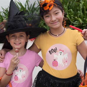 Puede incluir: Dos ni&ntilde;os vestidos con disfraces de Halloween. Un ni&ntilde;o lleva un sombrero de bruja negro y una camiseta rosa con un gr&aacute;fico de fantasma y el texto "BOO!" El otro ni&ntilde;o lleva una camiseta amarilla con un gr&aacute;fico de fantasma y el texto "BOO!"