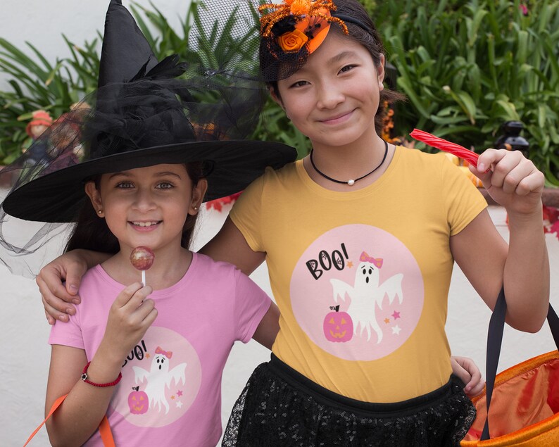 Puede incluir: Dos ni&ntilde;os vestidos con disfraces de Halloween. Un ni&ntilde;o lleva un sombrero de bruja negro y una camiseta rosa con un gr&aacute;fico de fantasma y el texto "BOO!" El otro ni&ntilde;o lleva una camiseta amarilla con un gr&aacute;fico de fantasma y el texto "BOO!"