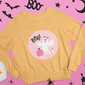Puede incluir: Una sudadera amarilla con un dise&ntilde;o circular rosa que presenta un fantasma con un lazo, una calabaza y el texto "BOO!"