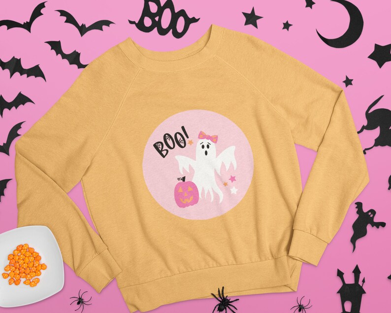 Puede incluir: Una sudadera amarilla con un dise&ntilde;o circular rosa que presenta un fantasma con un lazo, una calabaza y el texto "BOO!"
