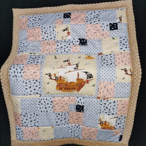 Peut inclure: Une courtepointe b&eacute;b&eacute; beige avec un motif patchwork repr&eacute;sentant un bateau, des ancres et d'autres motifs nautiques. La courtepointe est faite d'un tissu doux et confortable et est parfaite pour une chambre de b&eacute;b&eacute; ou une salle de jeux.