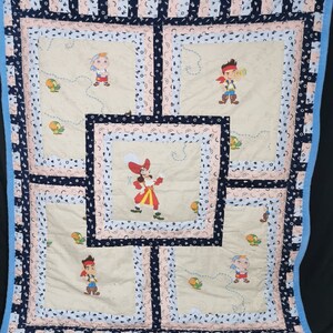 Könnte beinhalten: Eine beige gesteppte Decke mit einem blauen und rosa Rand. Die Decke hat ein sich wiederholendes Muster aus Sternen, Monden und Ankern. Auf der Decke sind fünf gestickte Bilder von Piraten zu sehen.