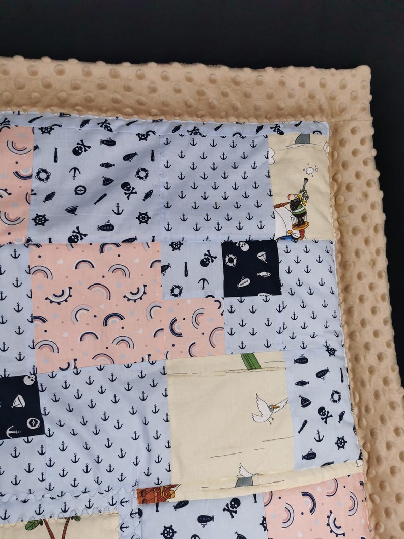 Peut inclure: Une couverture b&eacute;b&eacute; en patchwork sur le th&egrave;me nautique. La couverture est faite de diff&eacute;rents tissus avec des motifs vari&eacute;s, notamment des ancres, des arcs-en-ciel et un bateau pirate. La couverture est bord&eacute;e d'un tissu beige doux.