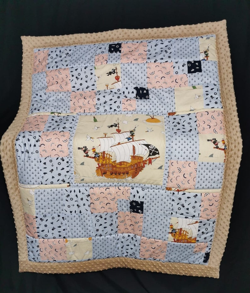 Peut inclure: Une courtepointe b&eacute;b&eacute; beige avec un motif patchwork repr&eacute;sentant un bateau, des ancres et d'autres motifs nautiques. La courtepointe est faite d'un tissu doux et confortable et est parfaite pour une chambre de b&eacute;b&eacute; ou une salle de jeux.